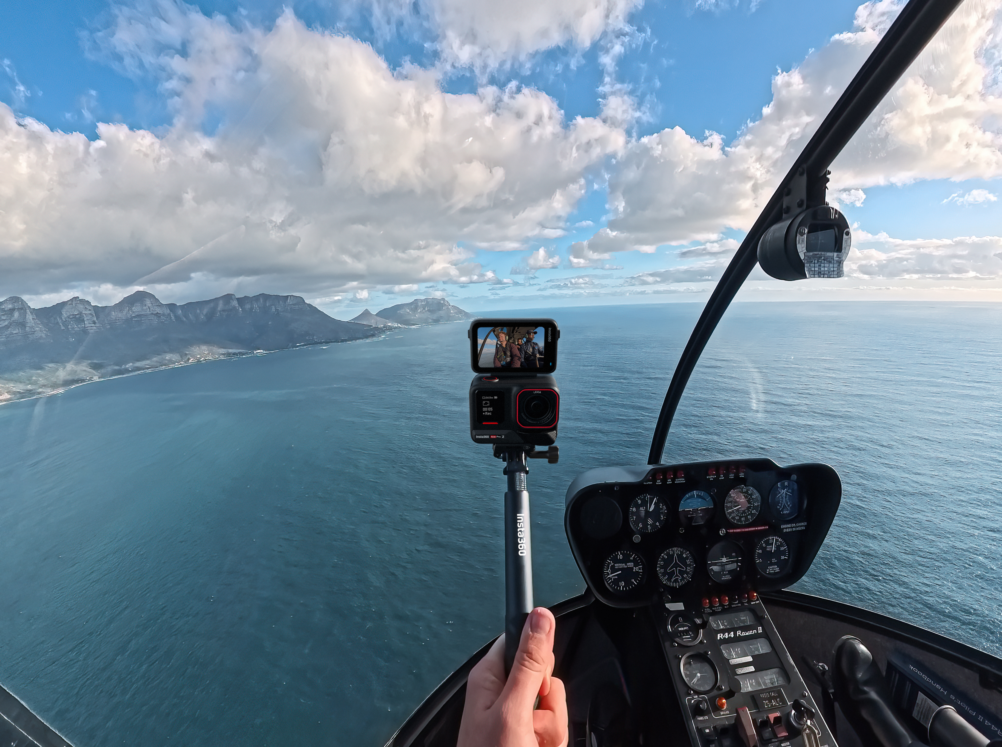 Insta360 Ace Pro 2 attached to a selfie stick with the flip screen open. The person is in a plane's cockpit flying over a scenic ocean.